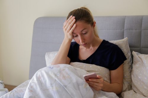 woman sitting on bed looking at her phone with hand on head looking shocked