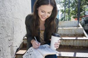 a person with long brown hair sitting outside drinking coffee and doing a crossword puzzle for better brain health