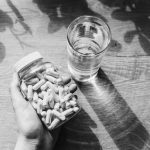 black and white photo of hand holding jar of supplement capsules next to glass of water
