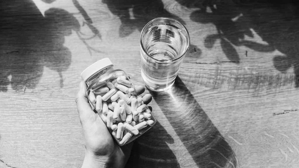 black and white photo of hand holding jar of supplement capsules next to glass of water