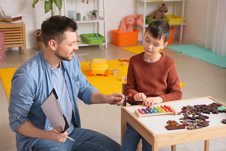 A boy and a man sit down at a table. The boy is playing a colorful xylophone.