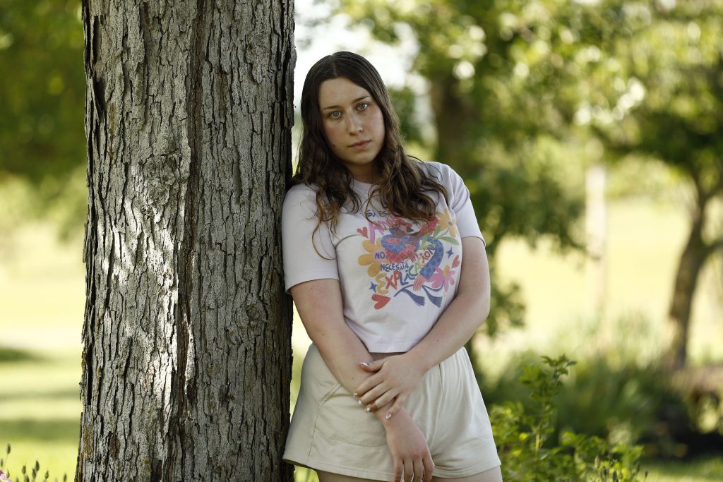 Gwen Schwarz is photographed standing outdoors on a sunny day. She leans against a tree and looks directly into the camera. She is wearing a colorful shirt and tan shorts.
