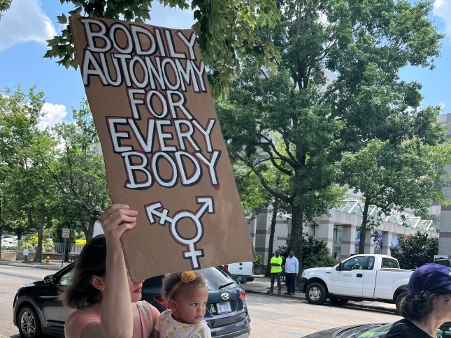 A woman holding a baby holds up a cardboard sign that reads "Body autonomy for each body."