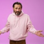 Close-up of a man in a light pink hoodie shrugging with arms outstretched