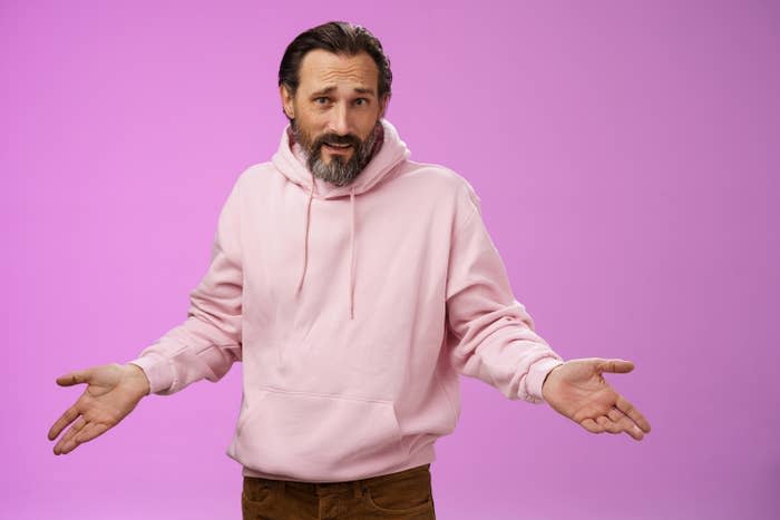 Close-up of a man in a light pink hoodie shrugging with arms outstretched
