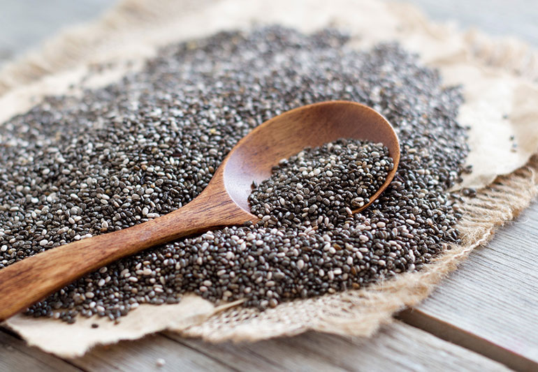 A wooden spoon scooping out some chia seeds from a pile of chia seeds in a burlap wrapper on a wooden table.