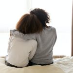 A woman with her arm around a teenager, sitting on the edge of a bed, seen from behind