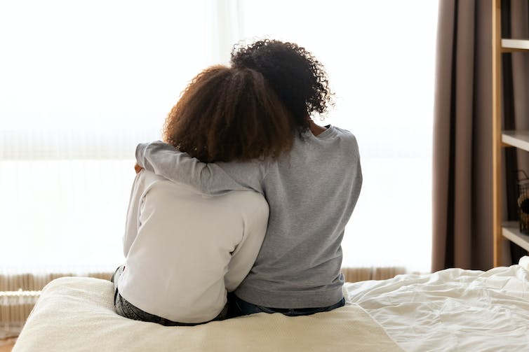 A woman with her arm around a teenager, sitting on the edge of a bed, seen from behind