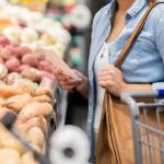 Woman holds shopping list and bag as she stands in front of baskets of potatoes in a supermarket.