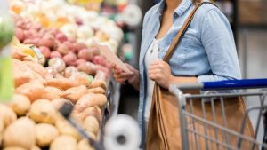 Woman holds shopping list and bag as she stands in front of baskets of potatoes in a supermarket.